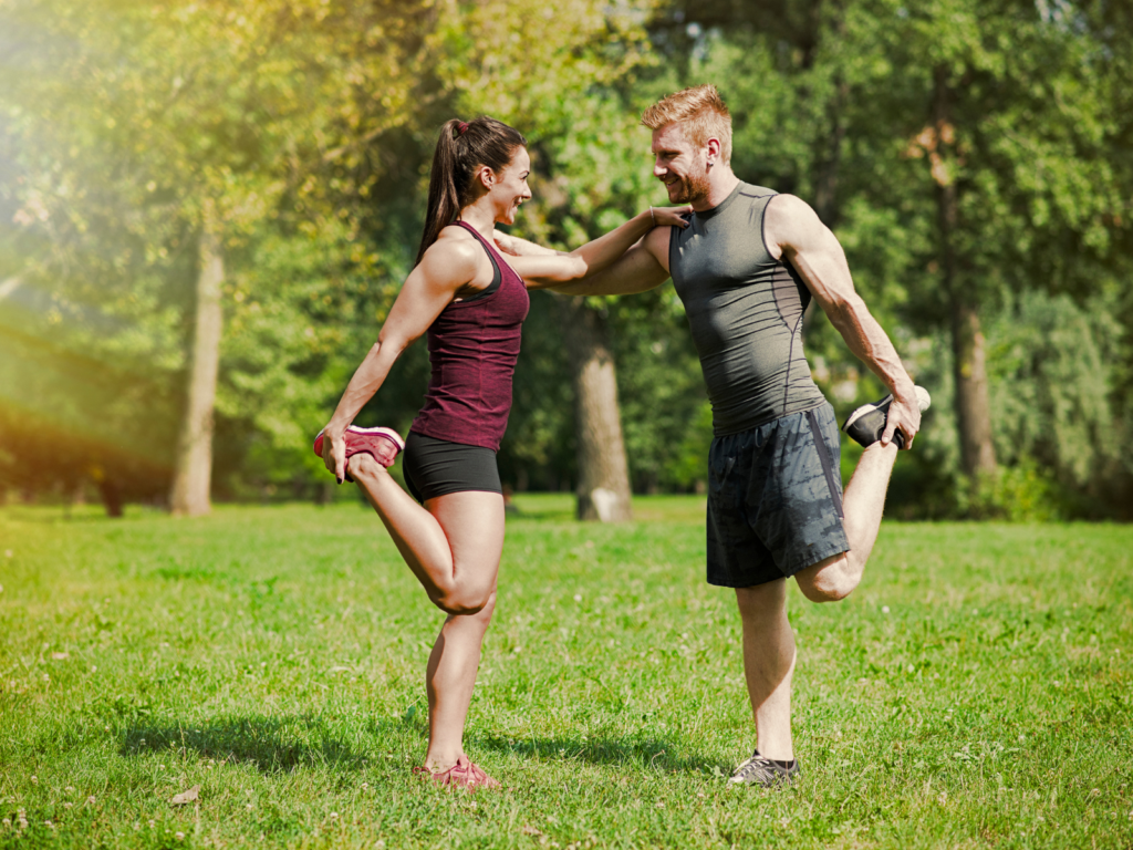 Casal que vão treinar juntos. Um casal sorridente, um homem e uma mulher, realiza um alongamento de quadríceps em pé em um parque gramado e ensolarado. Eles estão de frente um para o outro, cada um equilibrando-se em uma perna enquanto segura o pé oposto atrás do corpo. Eles apoiam as mãos nos ombros um do outro para manter o equilíbrio, trocando olhares cúmplices. O homem veste uma regata cinza e shorts escuros; a mulher usa uma regata vinho e shorts pretos. Ao fundo, há árvores verdes e uma iluminação suave de fim de tarde.