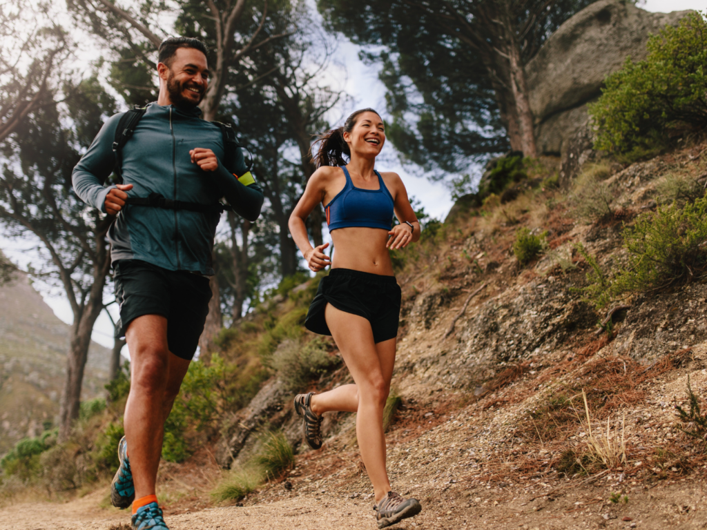 Um casal sorridente corre junto em uma trilha de terra em meio à natureza. A mulher, em primeiro plano à direita, usa um top esportivo azul, shorts pretos e tênis de trilha. O homem, ligeiramente atrás à esquerda, veste uma jaqueta esportiva cinza, shorts pretos e carrega uma mochila de hidratação. Ambos aparentam estar felizes e em sintonia. O cenário é composto por uma encosta inclinada com vegetação rasteira, árvores altas ao fundo e um céu claro. A foto é tirada de um ângulo baixo, reforçando a sensação de movimento e superação.